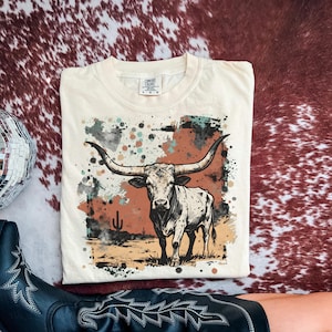 May include: Cream-colored t-shirt featuring a graphic of a longhorn steer with large horns, set against a desert landscape with a cactus. The design includes splashes of color in shades of brown, teal, and black. The shirt is a Comfort Colors brand.