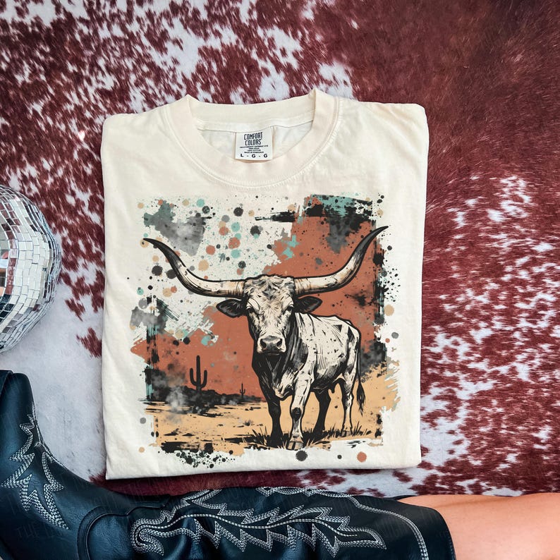 May include: Cream-colored t-shirt featuring a graphic of a longhorn steer with large horns, set against a desert landscape with a cactus. The design includes splashes of color in shades of brown, teal, and black. The shirt is a Comfort Colors brand.