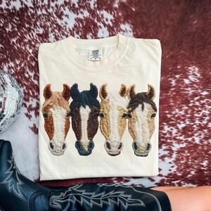 Peut inclure: T-shirt cr&egrave;me Comfort Colors avec un motif brod&eacute; de quatre t&ecirc;tes de chevaux dans les tons marron, noir et beige. Le t-shirt est pli&eacute; sur une surface &agrave; motif peau de vache. Le t-shirt porte une &eacute;tiquette Comfort Colors.