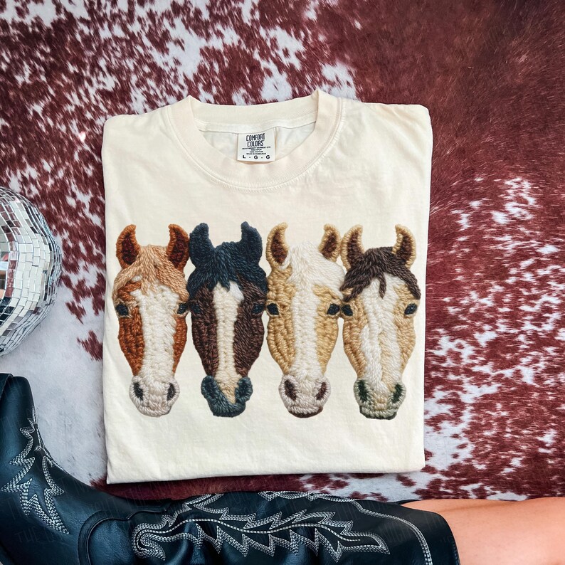 Peut inclure: T-shirt cr&egrave;me Comfort Colors avec un motif brod&eacute; de quatre t&ecirc;tes de chevaux dans les tons marron, noir et beige. Le t-shirt est pli&eacute; sur une surface &agrave; motif peau de vache. Le t-shirt porte une &eacute;tiquette Comfort Colors.