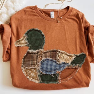 May include: A rust-colored t-shirt featuring a large, central duck-shaped appliqu&eacute;. The duck is constructed from various fabric patches, including dark green corduroy, brown plaid, blue gingham, and floral patterns with frayed, raw edges.