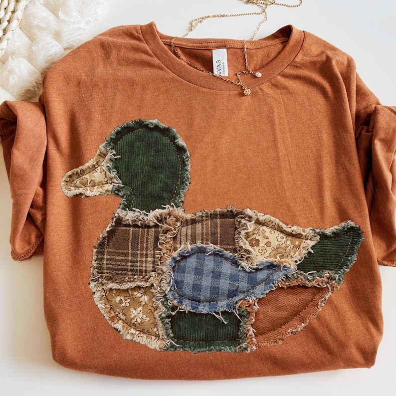 May include: A rust-colored t-shirt featuring a large, central duck-shaped appliqu&eacute;. The duck is constructed from various fabric patches, including dark green corduroy, brown plaid, blue gingham, and floral patterns with frayed, raw edges.