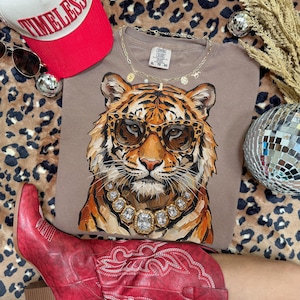 K&ouml;nnte beinhalten: Ein taupefarbenes T-Shirt mit einem Tiger, der eine Sonnenbrille und eine Diamantkette tr&auml;gt. Das Shirt wird mit einer roten Baseballkappe, Sonnenbrille und roten Cowboystiefeln kombiniert. Eine Discokugel und eine goldene Halskette sind ebenfalls zu sehen.