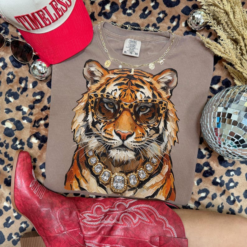 K&ouml;nnte beinhalten: Ein taupefarbenes T-Shirt mit einem Tiger, der eine Sonnenbrille und eine Diamantkette tr&auml;gt. Das Shirt wird mit einer roten Baseballkappe, Sonnenbrille und roten Cowboystiefeln kombiniert. Eine Discokugel und eine goldene Halskette sind ebenfalls zu sehen.