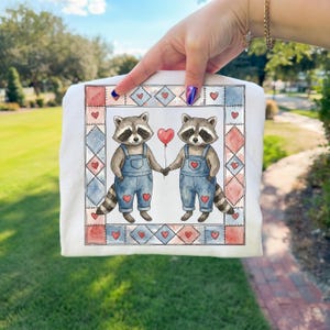 Pu&ograve; includere: Felpa bianca con un'illustrazione ad acquerello di due procioni che si tengono per mano e un palloncino rosso a forma di cuore. I procioni indossano salopette blu con dettagli a cuore. Il disegno &egrave; incorniciato da un bordo patchwork.