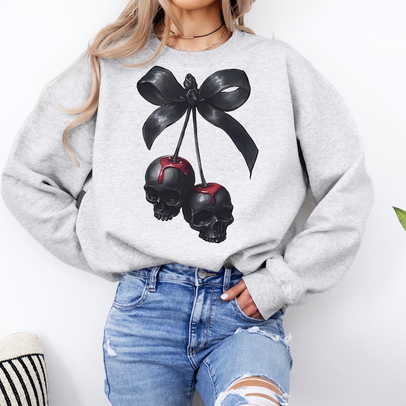 May include: A light grey sweatshirt with a graphic of two black skull cherries with red "juice" and a large black bow. The design is centered on the front of the sweatshirt. The model is wearing blue jeans.