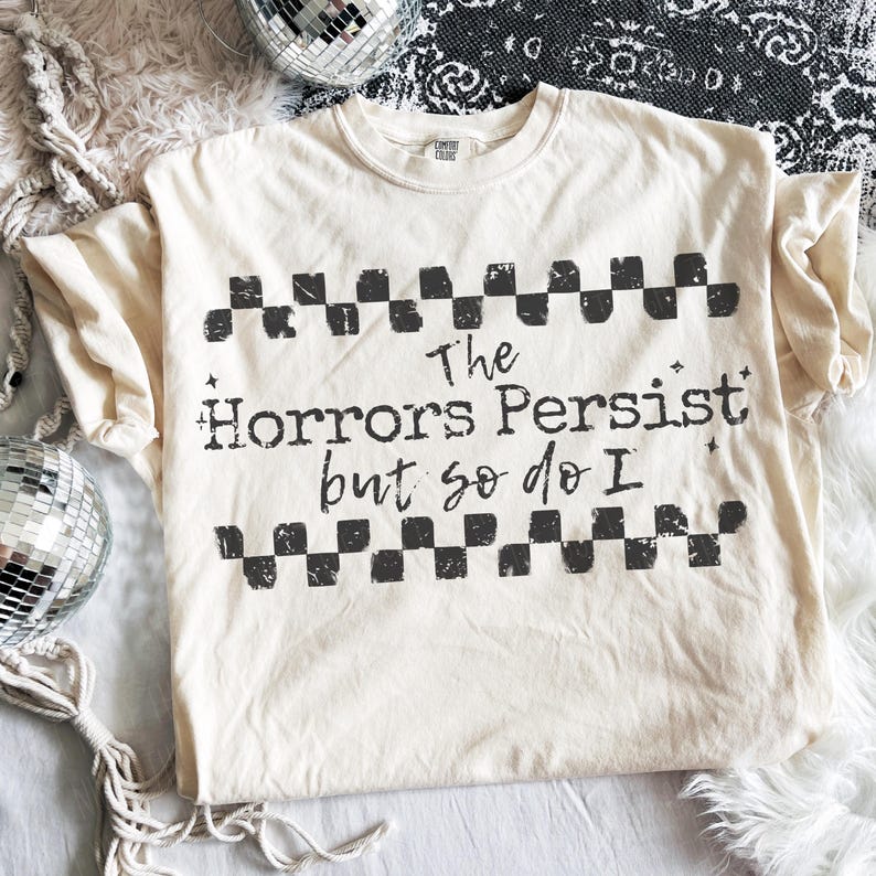 May include: Cream-colored t-shirt with the text "The Horrors Persist but so do I" in black, framed by a checkered pattern. The shirt is a Comfort Colors brand.