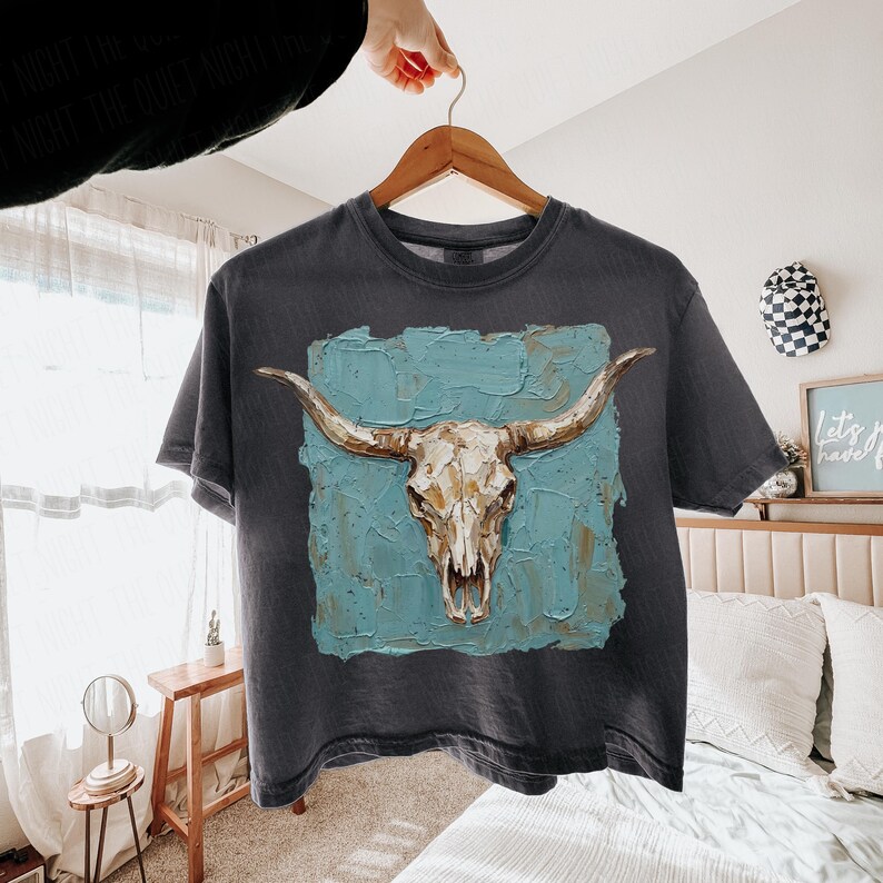 May include: Dark grey t-shirt with a bull skull illustration. The skull, with long horns, is set against a textured, turquoise background. The t-shirt is displayed on a wooden hanger.