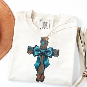 Peut inclure: T-shirt cr&egrave;me avec un motif de croix peinte et un n&oelig;ud bleu sarcelle. Un collier en or avec un pendentif repose sur le t-shirt. Le t-shirt porte une &eacute;tiquette Comfort Colors. Un chapeau marron et une plante s&eacute;ch&eacute;e sont en arri&egrave;re-plan.