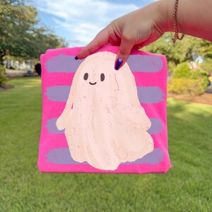 May include: A bright pink t-shirt featuring a central graphic of a pale peach-colored ghost with a simple smiling face. The ghost is layered over horizontal purple brushstroke stripes that span the width of the fabric.