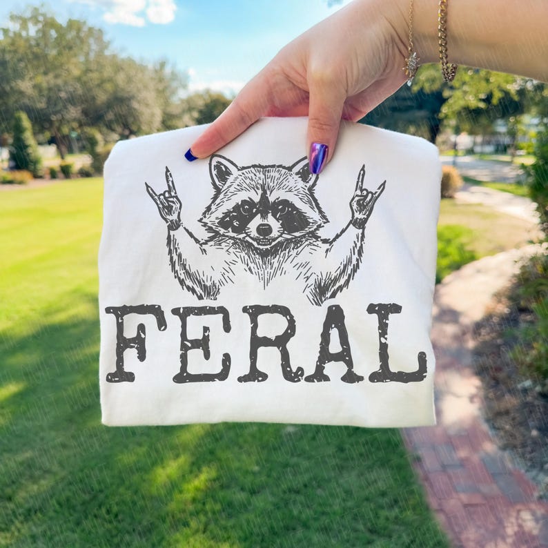 May include: White t-shirt featuring a black illustration of a raccoon making a rock and roll hand gesture. The word "FERAL" is printed in bold, black letters below the raccoon image.