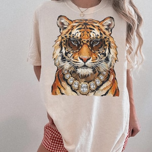 K&ouml;nnte beinhalten: Cremefarbenes T-Shirt mit einer detaillierten Illustration eines Tigergesichts. Der Tiger tr&auml;gt eine Sonnenbrille mit Leopardenmuster und eine Halskette mit Diamanten. Das Fell des Tigers ist orange und schwarz. Das Shirt hat kurze &Auml;rmel.