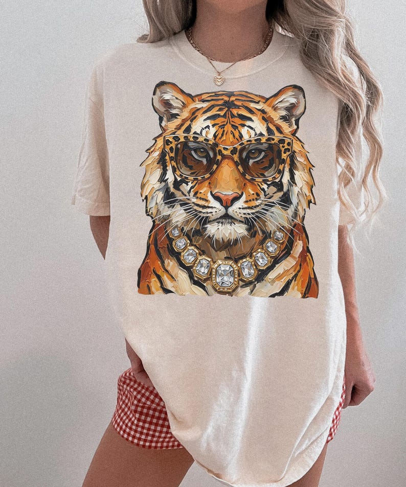 K&ouml;nnte beinhalten: Cremefarbenes T-Shirt mit einer detaillierten Illustration eines Tigergesichts. Der Tiger tr&auml;gt eine Sonnenbrille mit Leopardenmuster und eine Halskette mit Diamanten. Das Fell des Tigers ist orange und schwarz. Das Shirt hat kurze &Auml;rmel.