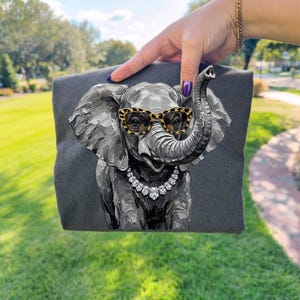 May include: A dark gray t-shirt featuring a textured, painterly illustration of an elephant. The elephant is depicted wearing leopard-print glasses and a silver-toned necklace with clear, faceted stones. The design has a thick, impasto style.