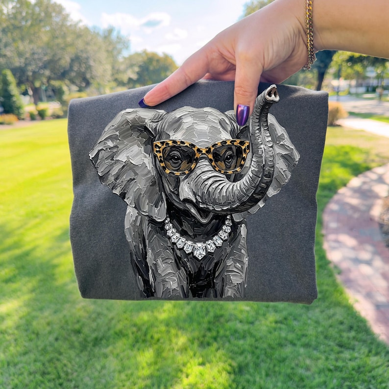 May include: A dark gray t-shirt featuring a textured, painterly illustration of an elephant. The elephant is depicted wearing leopard-print glasses and a silver-toned necklace with clear, faceted stones. The design has a thick, impasto style.
