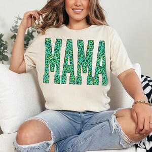 K&ouml;nnte beinhalten: Ein cremefarbenes Kurzarm-T-Shirt mit dem Wort MAMA in gro&szlig;en Blockbuchstaben. Die Buchstaben sind mit einem leuchtend gr&uuml;nen und gelben Leopardenmuster versehen, das einen auff&auml;lligen Kontrast zum hellen Stoff des Kleidungsst&uuml;cks bildet.