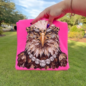 May include: A hot pink t-shirt featuring a detailed illustration of an eagle wearing leopard print glasses and a diamond necklace. The eagle's feathers are painted in shades of brown, beige, and white.
