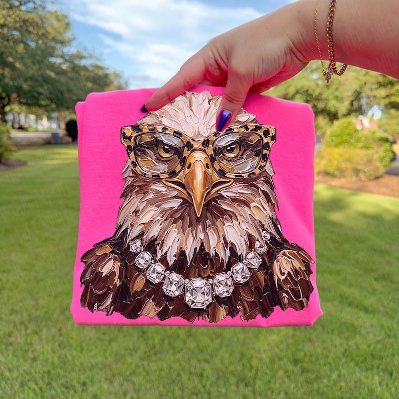 May include: A hot pink t-shirt featuring a detailed illustration of an eagle wearing leopard print glasses and a diamond necklace. The eagle's feathers are painted in shades of brown, beige, and white.
