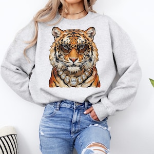 K&ouml;nnte beinhalten: Ein hellgraues Sweatshirt mit einer Aquarellillustration eines Tigers mit Sonnenbrille und Diamantkette. Der Tiger hat orange und schwarze Streifen. Das Sweatshirt ist ein l&auml;ssiger Stil.