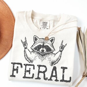 May include: Cream-colored t-shirt featuring a black graphic of a raccoon making a rock-and-roll hand gesture. The word "FERAL" is printed in bold, black letters below the raccoon. A gold necklace and a brown hat are also visible.
