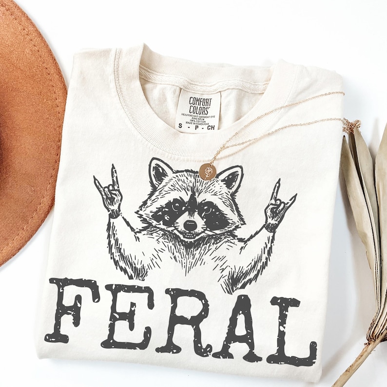 May include: Cream-colored t-shirt featuring a black graphic of a raccoon making a rock-and-roll hand gesture. The word "FERAL" is printed in bold, black letters below the raccoon. A gold necklace and a brown hat are also visible.