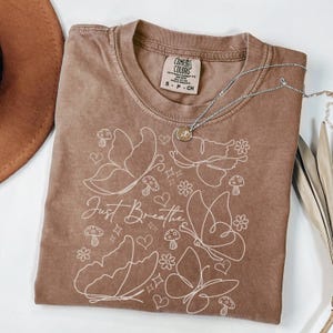 Puede incluir: Una camiseta marr&oacute;n Comfort Colors con un dise&ntilde;o de mariposas y flores blancas, y las palabras "Just Breathe". Un collar plateado con un colgante est&aacute; sobre la camiseta. La camiseta es talla S.