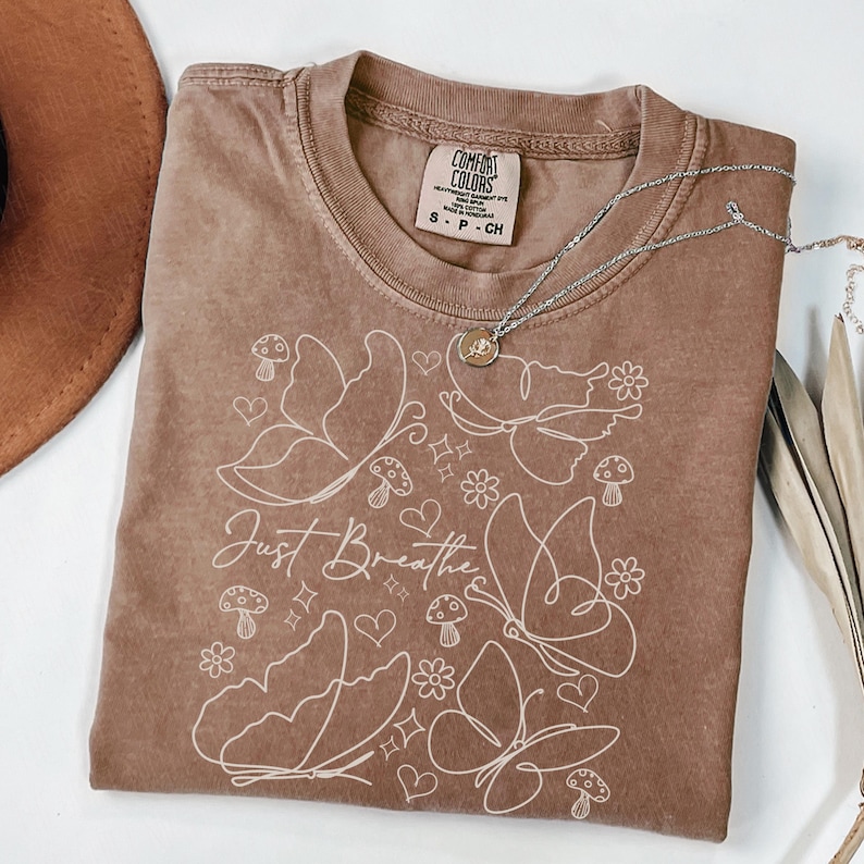 Puede incluir: Una camiseta marr&oacute;n Comfort Colors con un dise&ntilde;o de mariposas y flores blancas, y las palabras "Just Breathe". Un collar plateado con un colgante est&aacute; sobre la camiseta. La camiseta es talla S.