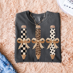 以下が含まれることがあります： 3つの装飾的な鉛筆とリボンが付いた濃い灰色のTシャツ。鉛筆は、白黒の市松模様、ヒョウ柄、黒い水玉模様の異なるパターンを持っています。各鉛筆には、タン色の消しゴムと鉛筆の先、タン色のリボンが付いています。