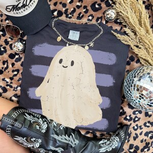 May include: A dark gray t-shirt featuring a beige ghost graphic with a cracked texture overlay, set against horizontal purple brush strokes. The shirt includes a gold-toned chain necklace with small charms resting on the collar area.