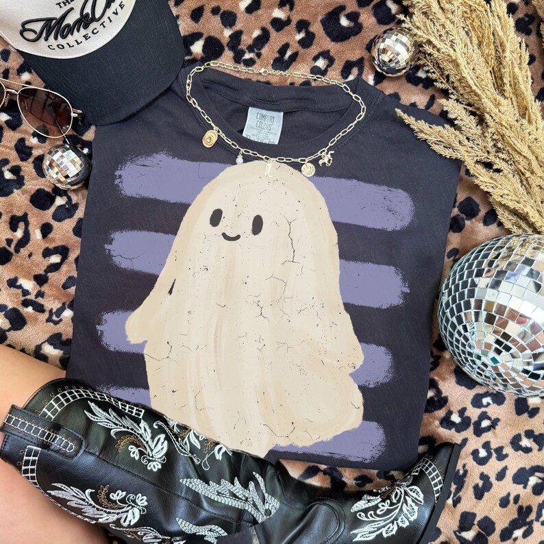 May include: A dark gray t-shirt featuring a beige ghost graphic with a cracked texture overlay, set against horizontal purple brush strokes. The shirt includes a gold-toned chain necklace with small charms resting on the collar area.