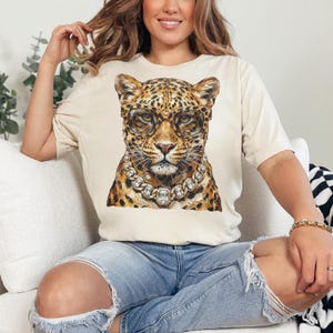 Puede incluir: Camiseta beige con una ilustraci&oacute;n de leopardo con gafas y un collar de diamantes. El leopardo tiene un pelaje manchado y est&aacute; centrado en la camiseta. La camiseta es de material suave y tiene mangas cortas.