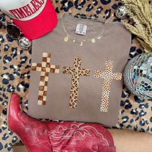 May include: A taupe-colored t-shirt featuring three crosses with different patterns: checkered, leopard print, and dotted. A red baseball cap with the word "TIMELESS" and a gold necklace with charms are also visible.