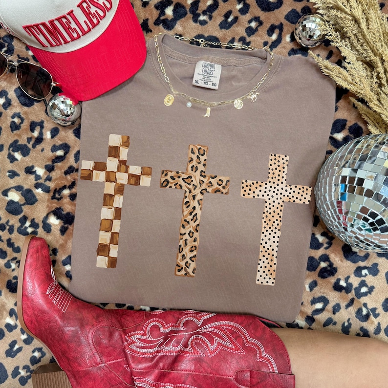 May include: A taupe-colored t-shirt featuring three crosses with different patterns: checkered, leopard print, and dotted. A red baseball cap with the word "TIMELESS" and a gold necklace with charms are also visible.