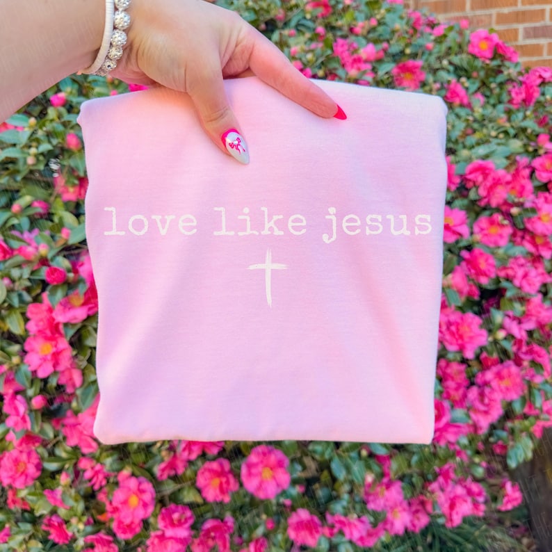 May include: A light pink sweatshirt with the words "love like jesus" and a cross printed in white. The sweatshirt is held up by a hand with a bracelet and nail art. The background is a bush with pink flowers.