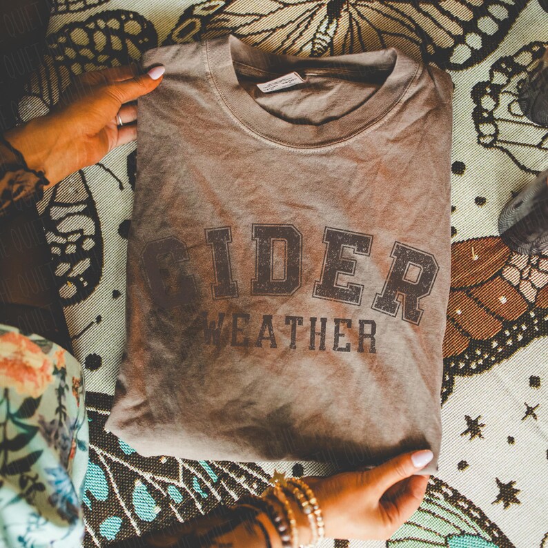 K&ouml;nnte beinhalten: Ein gefaltetes, braunes T-Shirt mit den Worten "CIDER WEATHER" in dunkelbraunen Blockbuchstaben aufgedruckt. Das Shirt hat einen Rundhalsausschnitt und ein Etikett auf der R&uuml;ckseite. Das Shirt wird von H&auml;nden mit Armb&auml;ndern und Ringen gehalten.