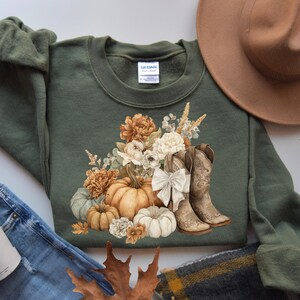 May include: An olive green sweatshirt featuring a fall-themed graphic. The design includes pumpkins, flowers, and cowboy boots. A brown hat and denim jeans are visible in the background. The sweatshirt has a crew neck and long sleeves.