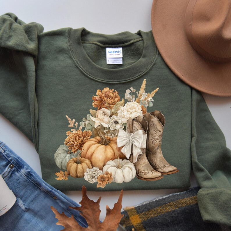 May include: An olive green sweatshirt featuring a fall-themed graphic. The design includes pumpkins, flowers, and cowboy boots. A brown hat and denim jeans are visible in the background. The sweatshirt has a crew neck and long sleeves.