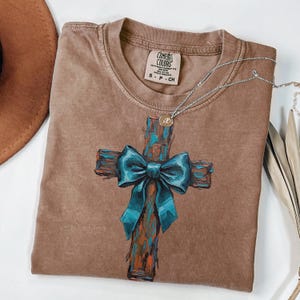 Peut inclure: Un t-shirt marron Comfort Colors avec une croix peinte et un n&oelig;ud bleu sarcelle. Le t-shirt a un col rond et des manches courtes. Un collier argent&eacute; avec un petit pendentif est drap&eacute; sur le t-shirt.