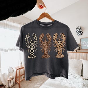May include: Dark gray t-shirt featuring three lobster designs. The lobsters have unique patterns: a black and white checkerboard, a leopard print, and a tan lobster with black spots. The shirt is hanging on a wooden hanger.