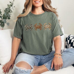 以下が含まれることがあります： オリーブグリーンのTシャツ。3匹のカニのデザインで、市松模様、ヒョウ柄、水玉模様が施されています。柔らかい素材で作られたTシャツです。