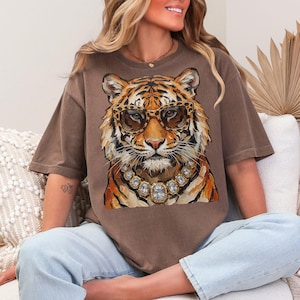 K&ouml;nnte beinhalten: Braunes T-Shirt mit einer Illustration eines Tigers, der eine Sonnenbrille und eine Diamantkette tr&auml;gt. Der Tiger hat orange und schwarze Streifen. Das T-Shirt ist locker geschnitten und hat kurze &Auml;rmel.
