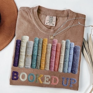 May include: A brown t-shirt with a crew neck featuring a colorful embroidered design of stacked books and the words "BOOKED UP" in a playful font. The shirt has a Comfort Colors tag and a silver necklace.