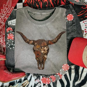 May include: A gray crew neck t-shirt featuring a detailed illustration of a longhorn skull adorned with floral accents in shades of brown, beige, and orange. The shirt is styled with denim shorts, a red cap, and cowboy boots, creating a western aesthetic.