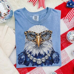 May include: A light blue t-shirt featuring a graphic of a bald eagle wearing leopard print glasses and a crystal-style necklace. The eagle is rendered in a textured, painterly style with white feathers and a yellow beak. The shirt has a Comfort Colors label.