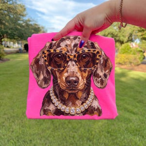 May include: A bright pink t-shirt featuring a graphic illustration of a dapple dachshund dog. The dog is wearing leopard print glasses and a chunky gold-toned chain necklace with clear square-cut stones. The artwork uses visible brushstrokes.