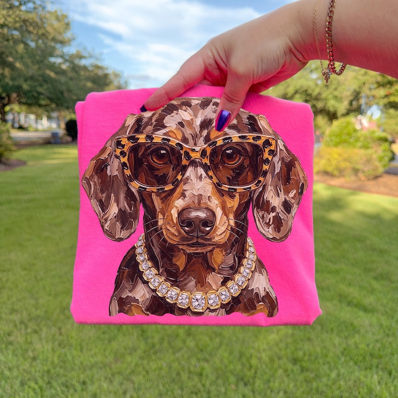 May include: A bright pink t-shirt featuring a graphic illustration of a dapple dachshund dog. The dog is wearing leopard print glasses and a chunky gold-toned chain necklace with clear square-cut stones. The artwork uses visible brushstrokes.