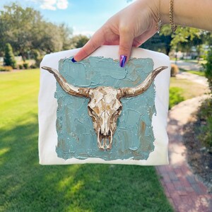 May include: White sweatshirt with a textured painting of a longhorn skull. The skull is a light brown colour with long, curved horns. The background is a square of blue-green paint.