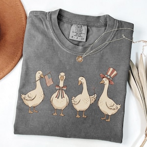 May include: Gray Comfort Colors t-shirt featuring four cartoon geese. One goose holds an American flag, another wears a bow tie, a third holds a sparkler, and the last wears a patriotic hat. A silver necklace with a pendant is also visible.