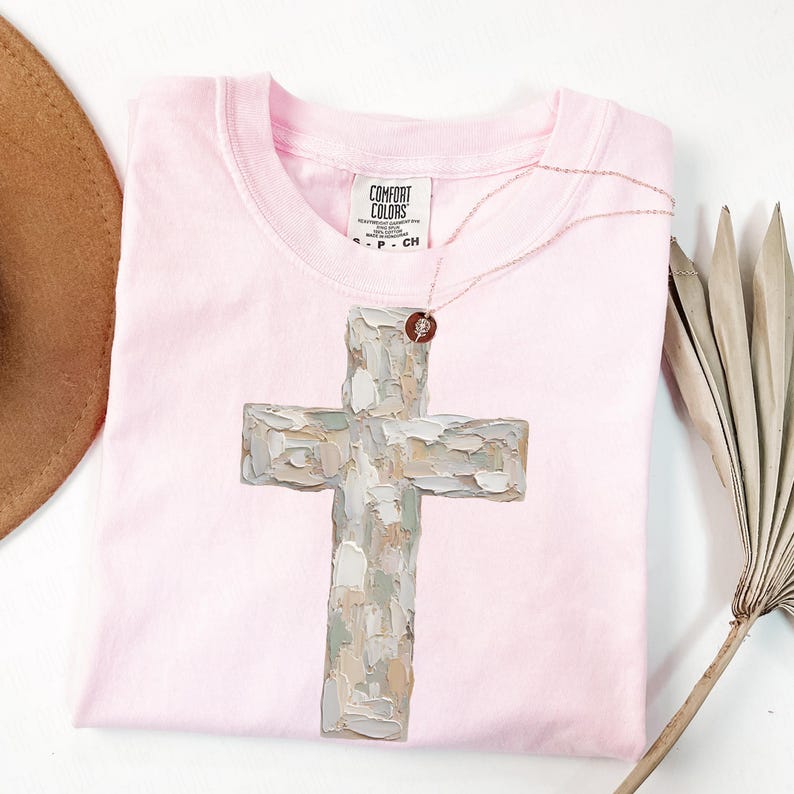 May include: A light pink t-shirt with a large, textured cross design in shades of gray, beige, and white. A delicate necklace with a small pendant rests on the shirt. The shirt is folded neatly, with a hat and dried plant in the background.