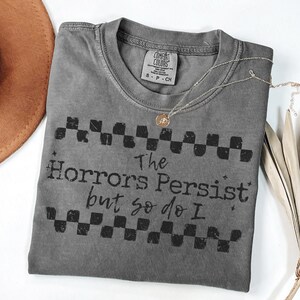 May include: A gray Comfort Colors t-shirt with the text "The Horrors Persist but so do I" in black. The shirt has a checkerboard design above and below the text. A gold necklace with a pendant and a brown hat are also visible.